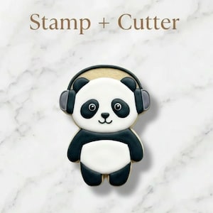 May include: A panda-shaped cookie cutter with a panda wearing headphones. The panda has black and white markings and a smiling face. The text "Stamp + Cutter" is at the top of the image. The cookie cutter is on a white marble background.