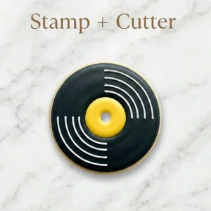 May include: A black and yellow cookie cutter in the shape of a vinyl record. The cookie has a black outer ring with a yellow center and white lines. The words "Stamp + Cutter" are at the top.