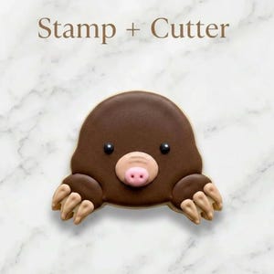 May include: A brown mole-shaped cookie cutter and stamp set. The cookie is decorated with a brown body, black eyes, a pink nose, and tan paws. The text "Stamp + Cutter" is at the top of the image.