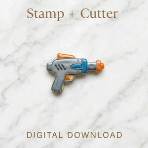 以下が含まれることがあります： グレー、ブルー、オレンジ色の光線銃のクッキーカッターとスタンプセット。クッキーカッターは未来的なブラスターの形をしています。「Stamp + Cutter」と「DIGITAL DOWNLOAD」の文字が表示されています。