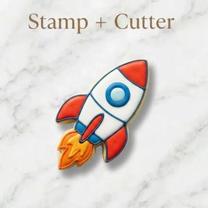May include: A rocket-shaped cookie cutter and stamp set featuring a white, red, and blue rocket design with a yellow and orange flame at the base. The text Stamp + Cutter is printed in brown above the rocket-shaped cookie.