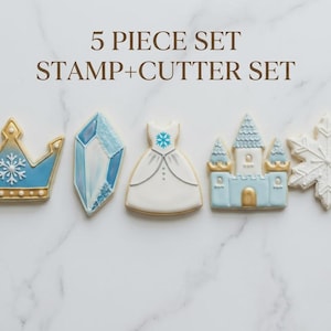 May include: A five-piece set of cookie cutters and stamps. The set includes a blue and gold crown, a blue gem, a white dress, a blue castle, and a white snowflake. The text "5 PIECE SET STAMP+CUTTER SET" is at the top.
