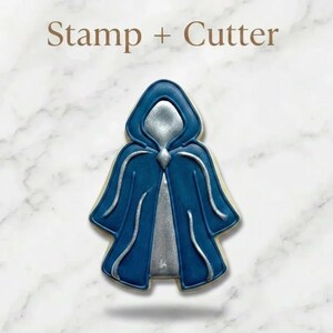 May include: A cookie cutter and stamp set shaped like a hooded cloak. The design features a deep blue outer cape with silver accents on the hood, trim, and inner garment. The text "Stamp + Cutter" is printed in brown above the cookie shape.