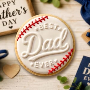 May include: A round sugar cookie decorated with white icing and red stitching to resemble a baseball. The cookie features the raised white text "BEST DAD EVER" in a script and block font style, centered on the surface of the cookie.