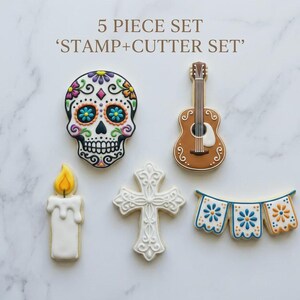 May include: A five-piece cookie cutter set featuring a sugar skull, guitar, candle, cross, and banner. The sugar skull is white with colorful floral details. The guitar is brown with white accents. The banner has blue and orange floral designs.