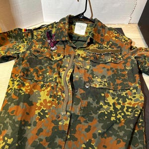Puede incluir: Chaqueta de camuflaje en tonos marrón, verde y amarillo. Cuenta con múltiples bolsillos, botones y un parche decorativo con un diseño de águila. La chaqueta está colgada en una percha negra. Diseño militar.