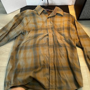May include: A long-sleeved button-down shirt in a plaid pattern of tan, brown, and grey. The shirt has a button-down collar, a chest pocket, and is hanging on a black hanger.