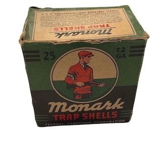 May include: Vintage Monark Trap Shells box. The box is green and tan with the words "Monark Trap Shells" in red and white. A circular graphic depicts a man in a red outfit holding a rifle. The box reads "25 12 GA" and "Federal Cartridge Corporation".