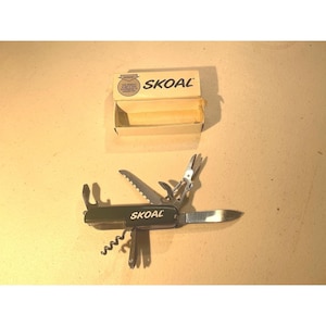 May include: A multi-tool with a black handle and the word "SKOAL" printed on it. The tool includes a knife blade, saw, scissors, and other implements. An open box with the word "SKOAL" is also in the image.