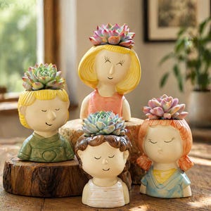 May include: Four ceramic succulent planters shaped like children's faces. Each planter has a different hair color and clothing color. The planters are filled with colorful succulents. The planters are sitting on a wooden surface.