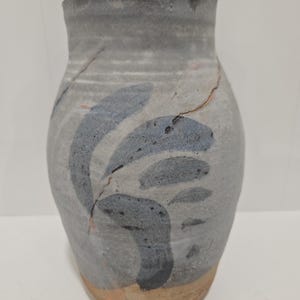 May include: A ceramic vase with a matte gray finish and a wide, rounded body. The surface features dark blue, brush-stroke leaf patterns and a visible crack running diagonally across the upper section. The base is unglazed, revealing a tan clay color.