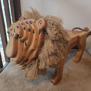 May include: A wooden bench shaped like a lion, featuring a carved light brown wood head with black painted spots, a thick mane made of natural tan fibers, four sturdy legs, and a short rope tail with a small red accent at the end of the tail.