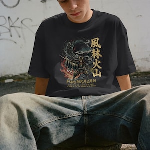 Puede incluir: Camiseta negra con un gráfico detallado de un guerrero samurái, texto japonés y las palabras "FURINKAZAN". El diseño incluye elementos de fuego, viento y un paisaje montañoso. La camiseta está hecha de un material suave.