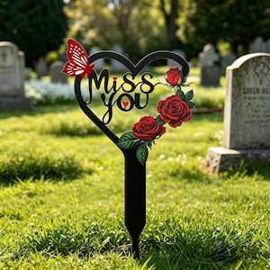 Pode incluir: Estaca de memorial em forma de coração de metal preto com as palavras "Miss You" e uma borboleta vermelha e rosas. A estaca é colocada em uma área gramada, provavelmente um cemitério, com lápides ao fundo.