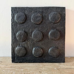 May include: A square, black wall art piece featuring a textured surface with nine raised, circular elements arranged in a three-by-three grid. The entire object has a matte, dark finish with a rough, tactile appearance across the flat base.