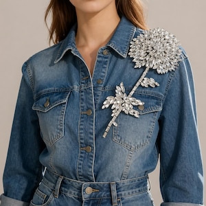 Puede incluir: Una camisa de mezclilla azul con una gran y compleja aplicaci&oacute;n floral en el hombro y el pecho izquierdos. La aplicaci&oacute;n est&aacute; compuesta por numerosos cristales transparentes facetados dispuestos en un patr&oacute;n de flor y tallo sobre la tela.