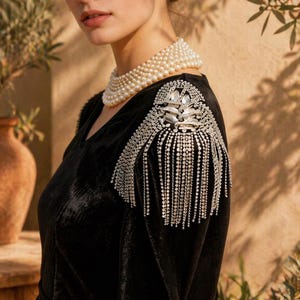 May include: A black velvet dress with a V-neck and embellished shoulder detail. The shoulder features a silver brooch with crystal fringe. A multi-strand pearl necklace adds elegance. The dress is a classic evening wear.