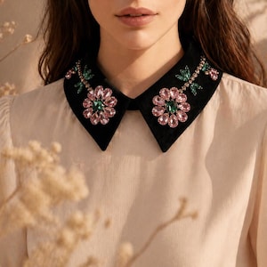 May include: A cream blouse featuring a black velvet collar decorated with pink and green floral detailing. The blouse has a round neckline and long sleeves. The collar is the focal point of the garment.