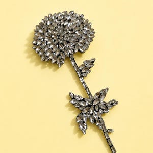 Puede incluir: Un broche con forma de flor elaborado con numerosos cristales transparentes de talla marquesa. El dise&ntilde;o presenta una gran cabeza floral circular y un tallo delgado adornado con hojas incrustadas de cristales sobre un fondo amarillo claro.