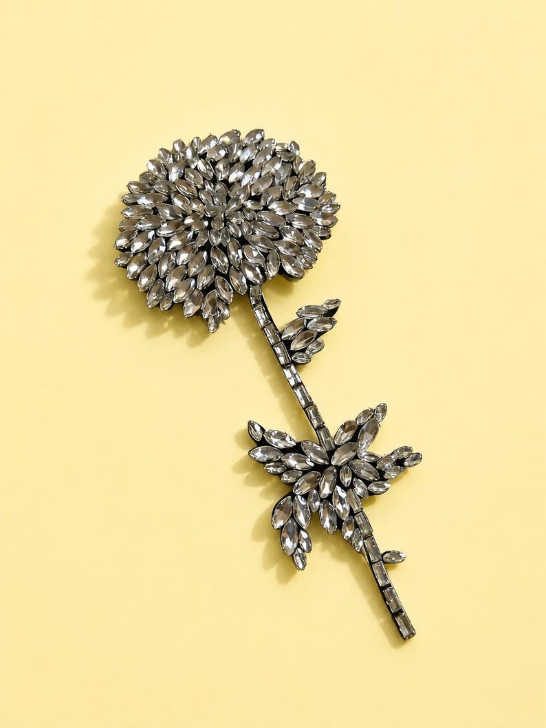 Puede incluir: Un broche con forma de flor elaborado con numerosos cristales transparentes de talla marquesa. El dise&ntilde;o presenta una gran cabeza floral circular y un tallo delgado adornado con hojas incrustadas de cristales sobre un fondo amarillo claro.