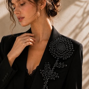 Puede incluir: Un blazer negro con una gran aplicaci&oacute;n floral intrincada en la solapa izquierda. El dise&ntilde;o de la flor est&aacute; elaborado con numerosas cuentas y cristales oscuros facetados, creando una apariencia texturizada y tridimensional en la tela.