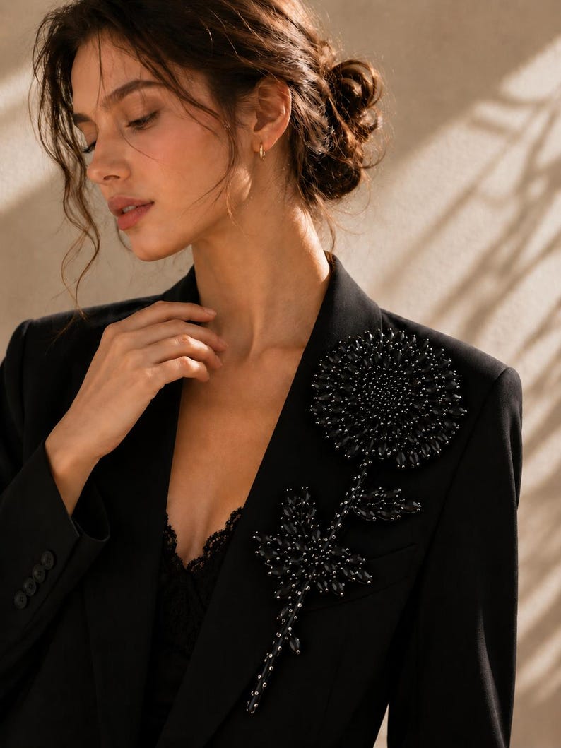 Puede incluir: Un blazer negro con una gran aplicaci&oacute;n floral intrincada en la solapa izquierda. El dise&ntilde;o de la flor est&aacute; elaborado con numerosas cuentas y cristales oscuros facetados, creando una apariencia texturizada y tridimensional en la tela.
