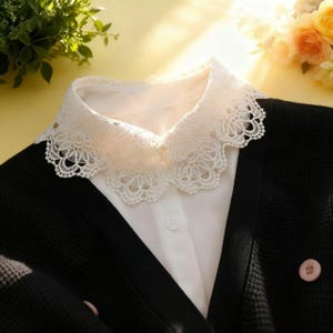 May include: A white lace collar over a white button-down shirt, paired with a black cardigan. The collar features a delicate, scalloped edge. The cardigan has pink buttons.