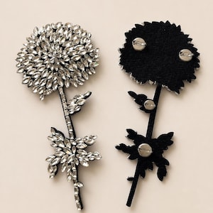 Puede incluir: Dos broches en forma de flor con cristales transparentes de talla marquesa dispuestos en un patr&oacute;n denso y superpuesto. Un broche muestra el frente incrustado de cristales, mientras que el otro muestra el reverso de fieltro negro con tres alfileres plateados.
