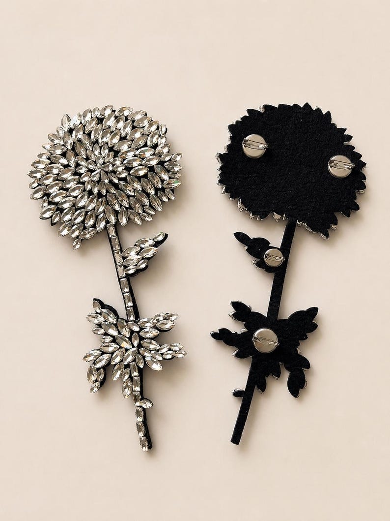 Puede incluir: Dos broches en forma de flor con cristales transparentes de talla marquesa dispuestos en un patr&oacute;n denso y superpuesto. Un broche muestra el frente incrustado de cristales, mientras que el otro muestra el reverso de fieltro negro con tres alfileres plateados.