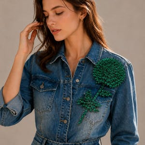 Puede incluir: Una camisa de mezclilla azul con botones y una gran aplicaci&oacute;n floral de cuentas de color verde oscuro en el pecho izquierdo. La camisa tiene un cuello cl&aacute;sico, mangas largas con pu&ntilde;os doblados y botones met&aacute;licos en la parte frontal.