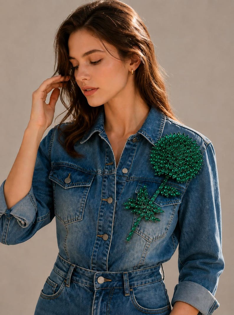 Puede incluir: Una camisa de mezclilla azul con botones y una gran aplicaci&oacute;n floral de cuentas de color verde oscuro en el pecho izquierdo. La camisa tiene un cuello cl&aacute;sico, mangas largas con pu&ntilde;os doblados y botones met&aacute;licos en la parte frontal.