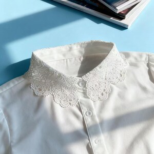 May include: A white blouse with a delicate, ornate lace collar. The collar is the focal point, featuring a detailed floral pattern. The blouse has a button-down front and is made of a smooth, likely cotton, fabric. The background is a light blue surface.