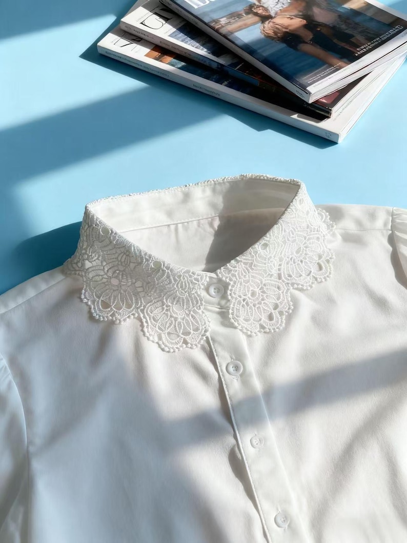 May include: A white blouse with a delicate, ornate lace collar. The collar is the focal point, featuring a detailed floral pattern. The blouse has a button-down front and is made of a smooth, likely cotton, fabric. The background is a light blue surface.