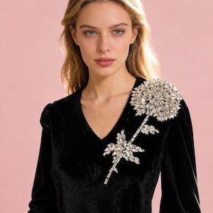 Puede incluir: Un vestido negro de terciopelo de manga larga con escote en V y una gran aplicaci&oacute;n floral en el hombro izquierdo. La flor est&aacute; construida con numerosos cristales transparentes facetados dispuestos en un denso patr&oacute;n de p&eacute;talos radiantes.