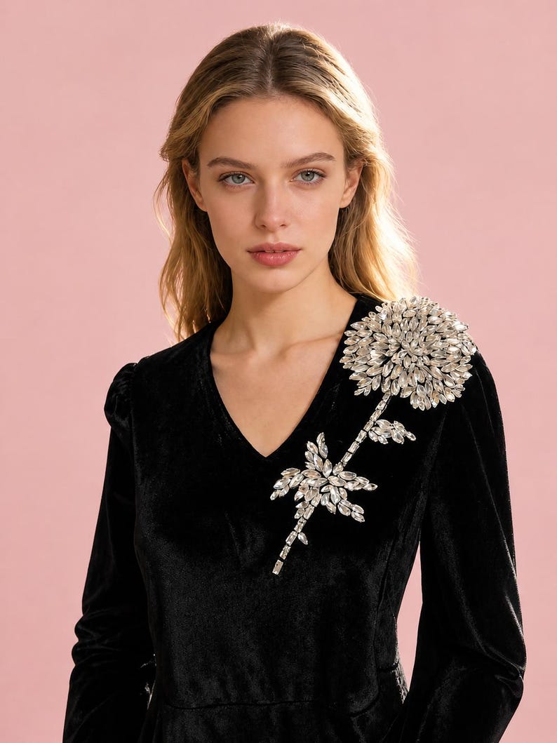 Puede incluir: Un vestido negro de terciopelo de manga larga con escote en V y una gran aplicaci&oacute;n floral en el hombro izquierdo. La flor est&aacute; construida con numerosos cristales transparentes facetados dispuestos en un denso patr&oacute;n de p&eacute;talos radiantes.