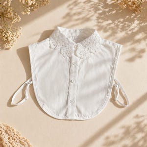 May include: A white detachable collar with a delicate lace trim. The collar has a button-down front and tie closures on each side. The collar is made of a crisp, white fabric and is a versatile accessory.