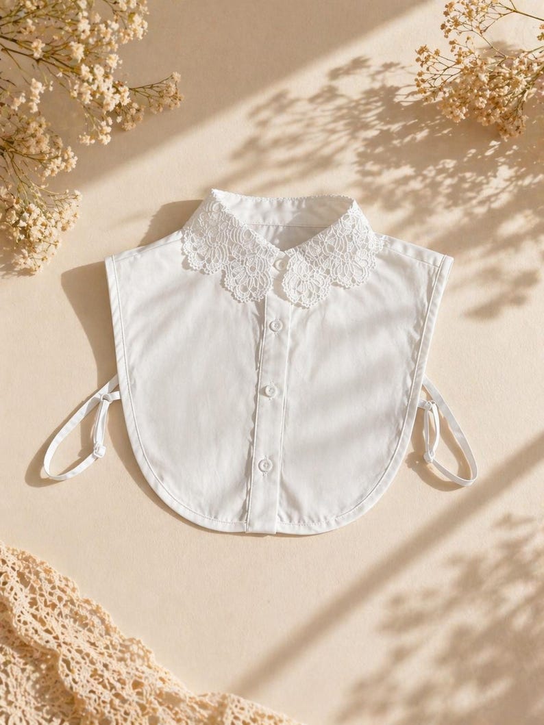May include: A white detachable collar with a delicate lace trim. The collar has a button-down front and tie closures on each side. The collar is made of a crisp, white fabric and is a versatile accessory.