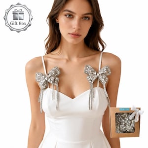 May include: A white dress with sparkling silver bow-shaped shoulder accessories. Each bow has dangling crystal strands. A gift box with similar accessories is also visible, suggesting a bridal or special occasion theme.
