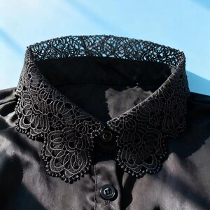 May include: Black satin shirt with a decorative lace collar. The collar features an intricate floral lace design. The shirt has a button-down front and a classic collar shape. The background is a light blue.