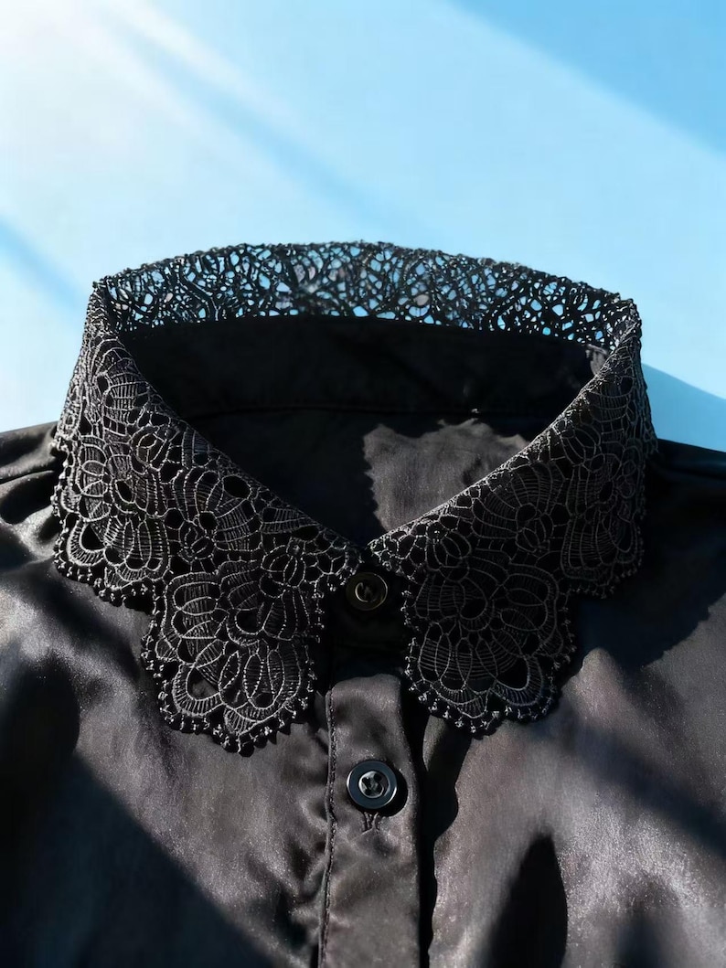 May include: Black satin shirt with a decorative lace collar. The collar features an intricate floral lace design. The shirt has a button-down front and a classic collar shape. The background is a light blue.