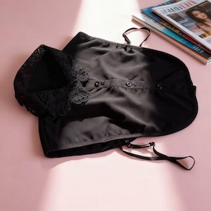 May include: Black sleepwear set with lace trim on the top and adjustable straps. The set includes a top with buttons and a matching bottom. The sleepwear is displayed on a pink surface with magazines in the background.