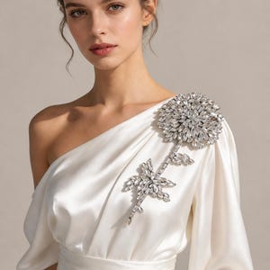 Puede incluir: Un vestido de sat&eacute;n color marfil de un solo hombro con una gran y compleja aplicaci&oacute;n floral en el hombro izquierdo. La aplicaci&oacute;n est&aacute; compuesta por numerosos cristales transparentes facetados en forma de flor y tallo en el corpi&ntilde;o.