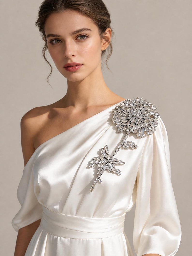 Puede incluir: Un vestido de sat&eacute;n color marfil de un solo hombro con una gran y compleja aplicaci&oacute;n floral en el hombro izquierdo. La aplicaci&oacute;n est&aacute; compuesta por numerosos cristales transparentes facetados en forma de flor y tallo en el corpi&ntilde;o.