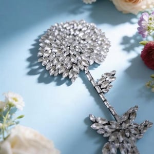 Puede incluir: Un broche o aplique decorativo en forma de flor elaborado con numerosos cristales transparentes de talla marquesa dispuestos en una cabeza circular densa, con un tallo delgado que presenta hojas de cristal m&aacute;s peque&ntilde;as y brillantes.