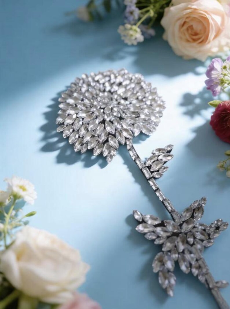 Puede incluir: Un broche o aplique decorativo en forma de flor elaborado con numerosos cristales transparentes de talla marquesa dispuestos en una cabeza circular densa, con un tallo delgado que presenta hojas de cristal m&aacute;s peque&ntilde;as y brillantes.