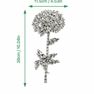Puede incluir: Un adorno floral de tono plateado incrustado con cristales transparentes, que presenta una gran flor circular y un tallo con hojas. El art&iacute;culo mide 11,5 cm de ancho y 26 cm de alto, seg&uacute;n lo indicado por las marcas de medici&oacute;n visibles.