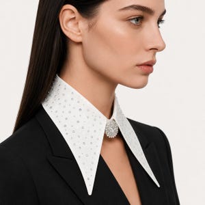 May include: A detachable white pointed collar featuring an all-over pattern of small, clear, reflective crystals. The collar is secured at the center front with a large, circular, multifaceted crystal brooch, worn over a solid black blazer.