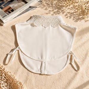 May include: A white detachable collar with a delicate lace trim. The collar has a rounded shape and tie closures on each side. The item is displayed on a neutral-toned surface with a magazine in the background.