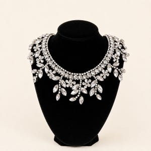 May include: A statement necklace featuring a wide collar of clear, round-cut rhinestones. The lower edge is adorned with a series of dangling floral and leaf-shaped crystal accents, creating a layered, botanical-inspired design in silver-tone metal.