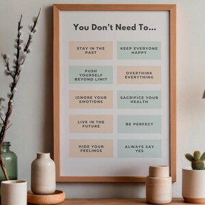 May include: A framed print with the text "You Don't Need To..." and a list of statements in a minimalist design. The print is in a light wood frame, and the background is a neutral color. Decorative items like a vase with branches and a cactus are in the foreground.